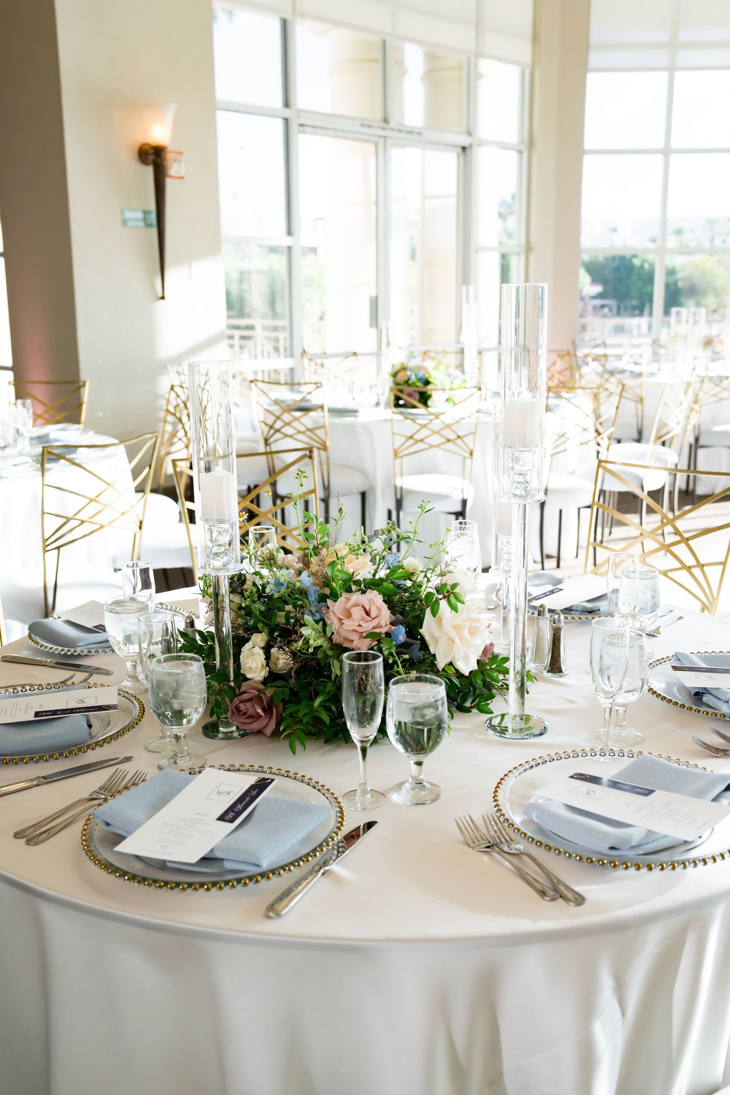 Elegant wedding reception table with floral centerpiece, gold chargers, gray napkins, and candles in a bright venue.