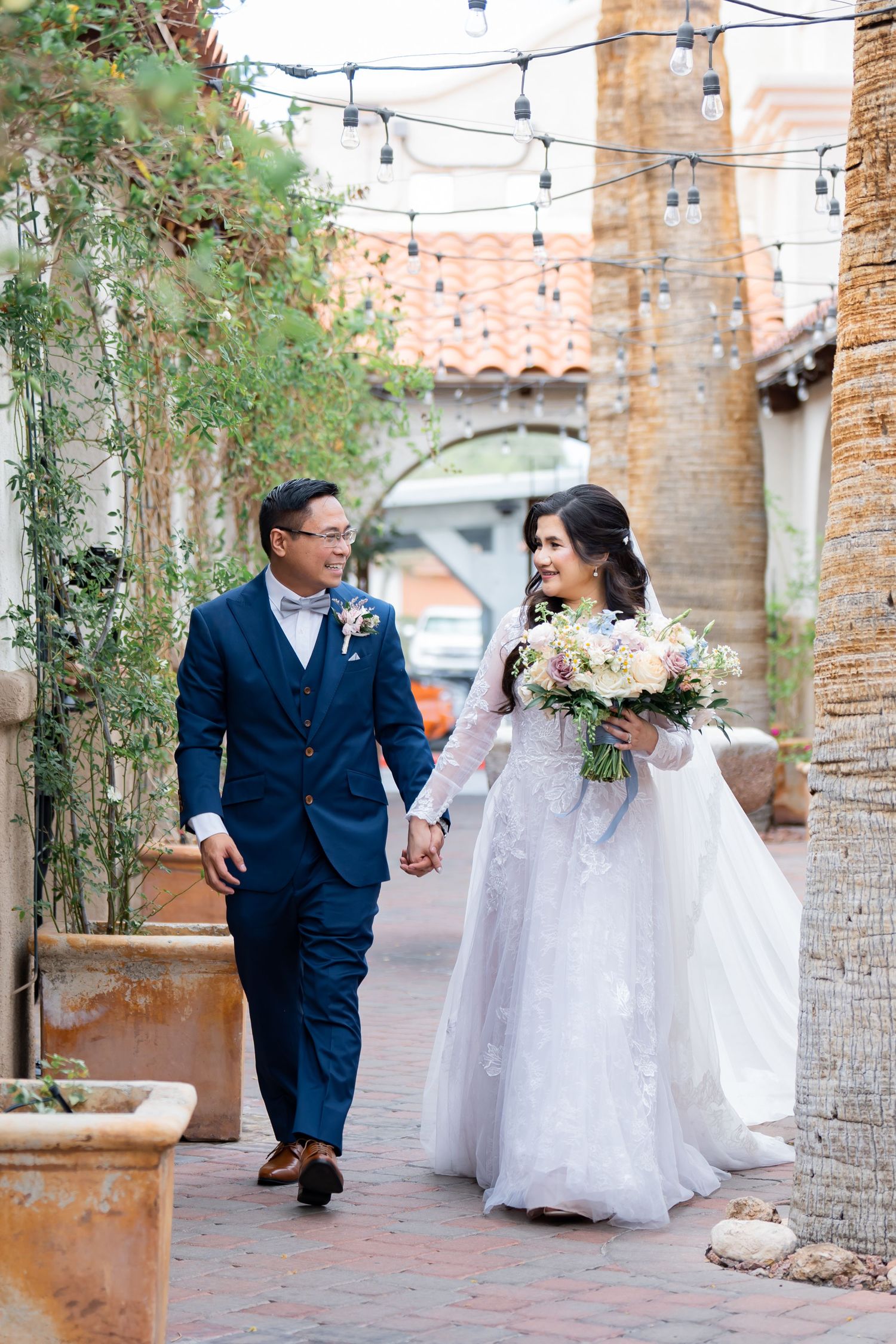 A bride and groom walk hand-in-hand through a charming courtyard, she in a lace gown holding a bouquet, he in a navy suit.