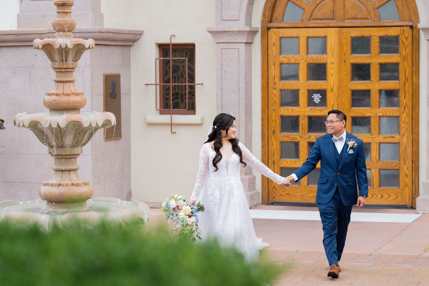 Bride and groom holding hands walking outside a elegant building with a stone fountain on their wedding day.
