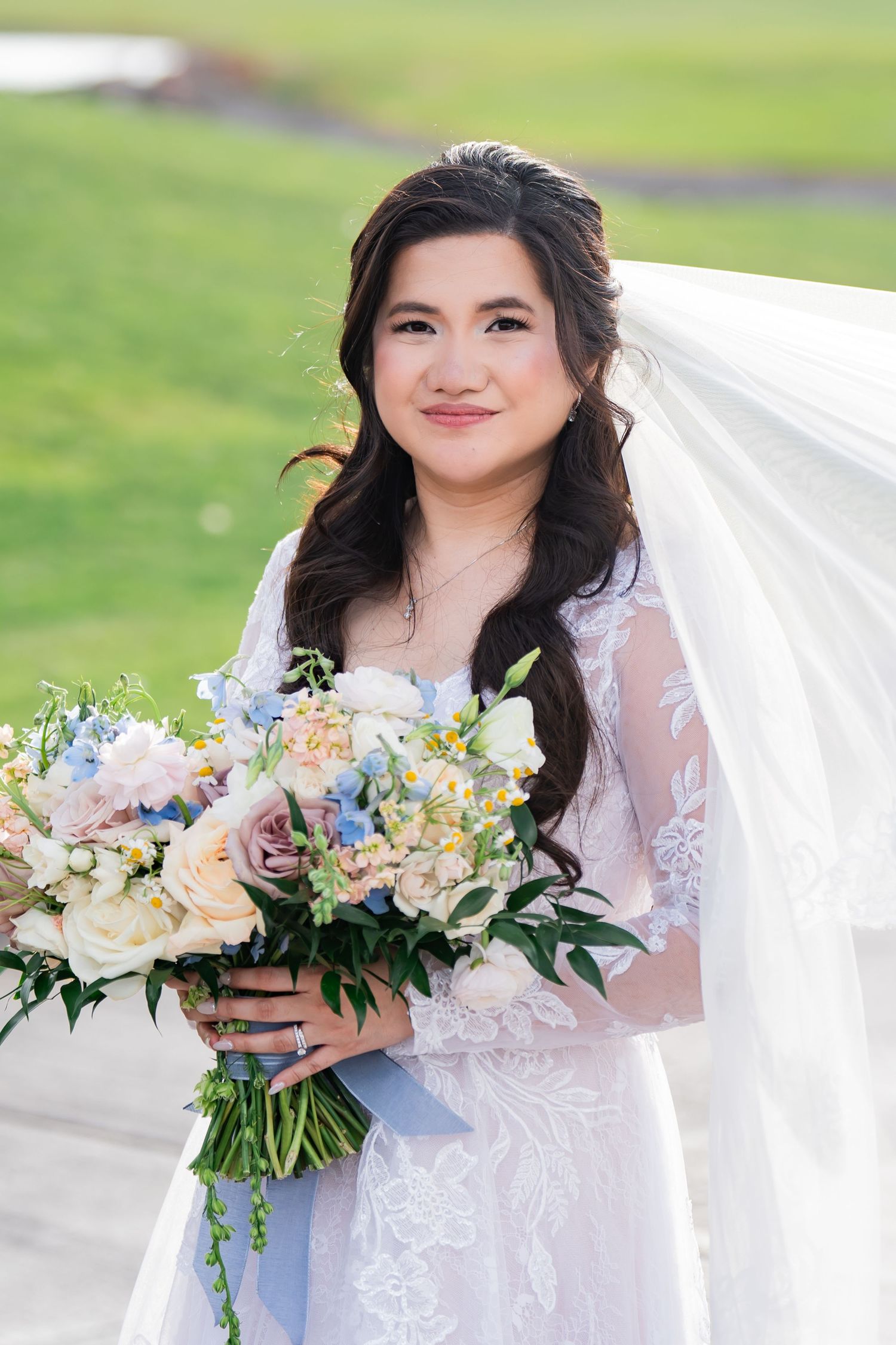 Smiling bride in lace gown holds pastel bouquet of roses and hydrangeas, veil flowing, outdoors on wedding day.