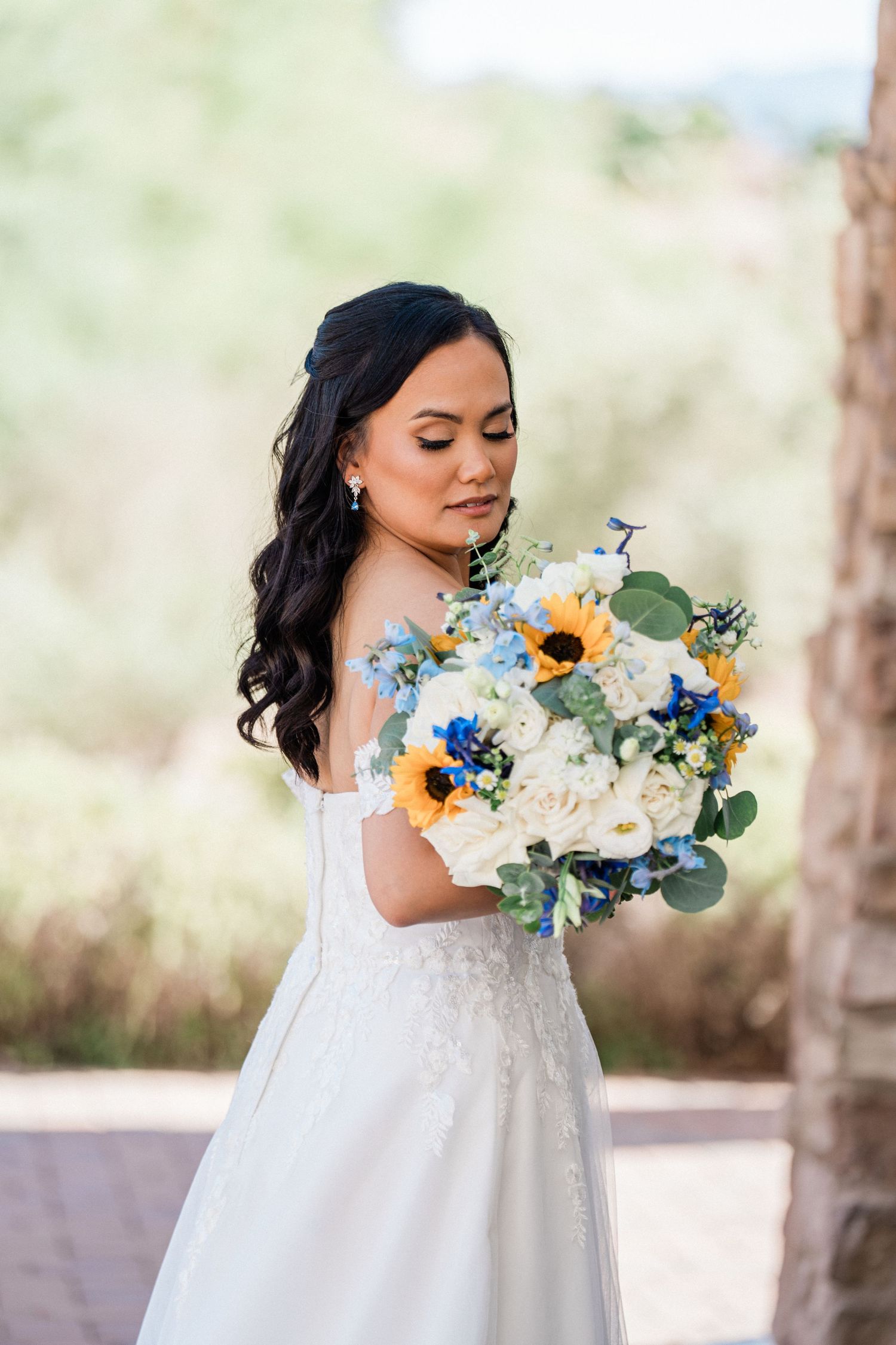 Bride in white off-shoulder gown holding sunflower and white rose bouquet with blue accents outdoors.