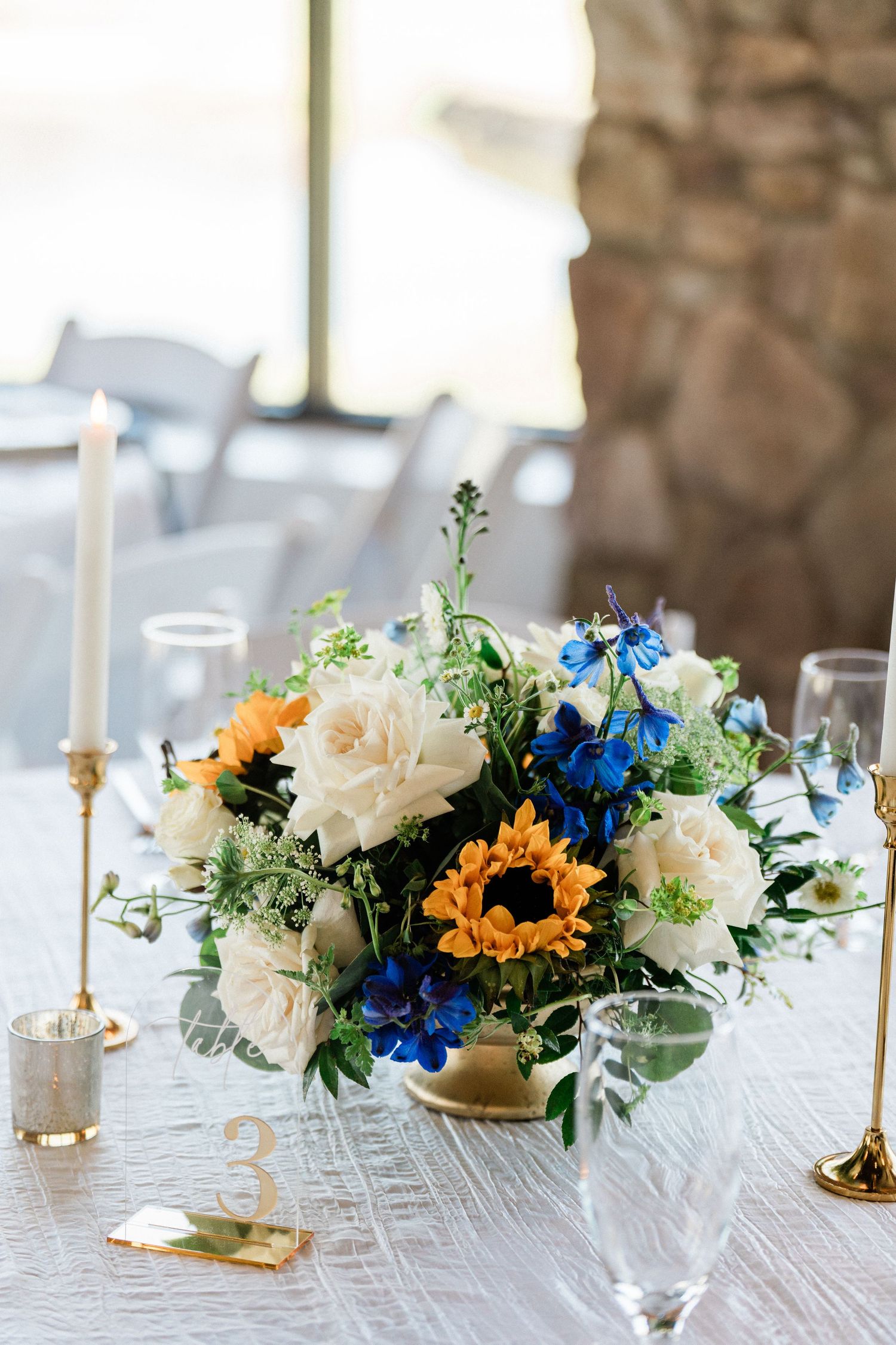 Blue and white wedding centerpiece with sunflowers, roses, and greenery in a gold vase on table number 3.