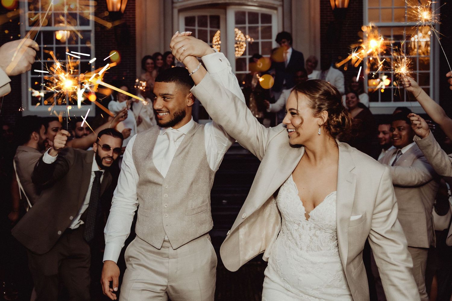 Newlyweds in matching white outfits dancing together while guests hold sparklers.