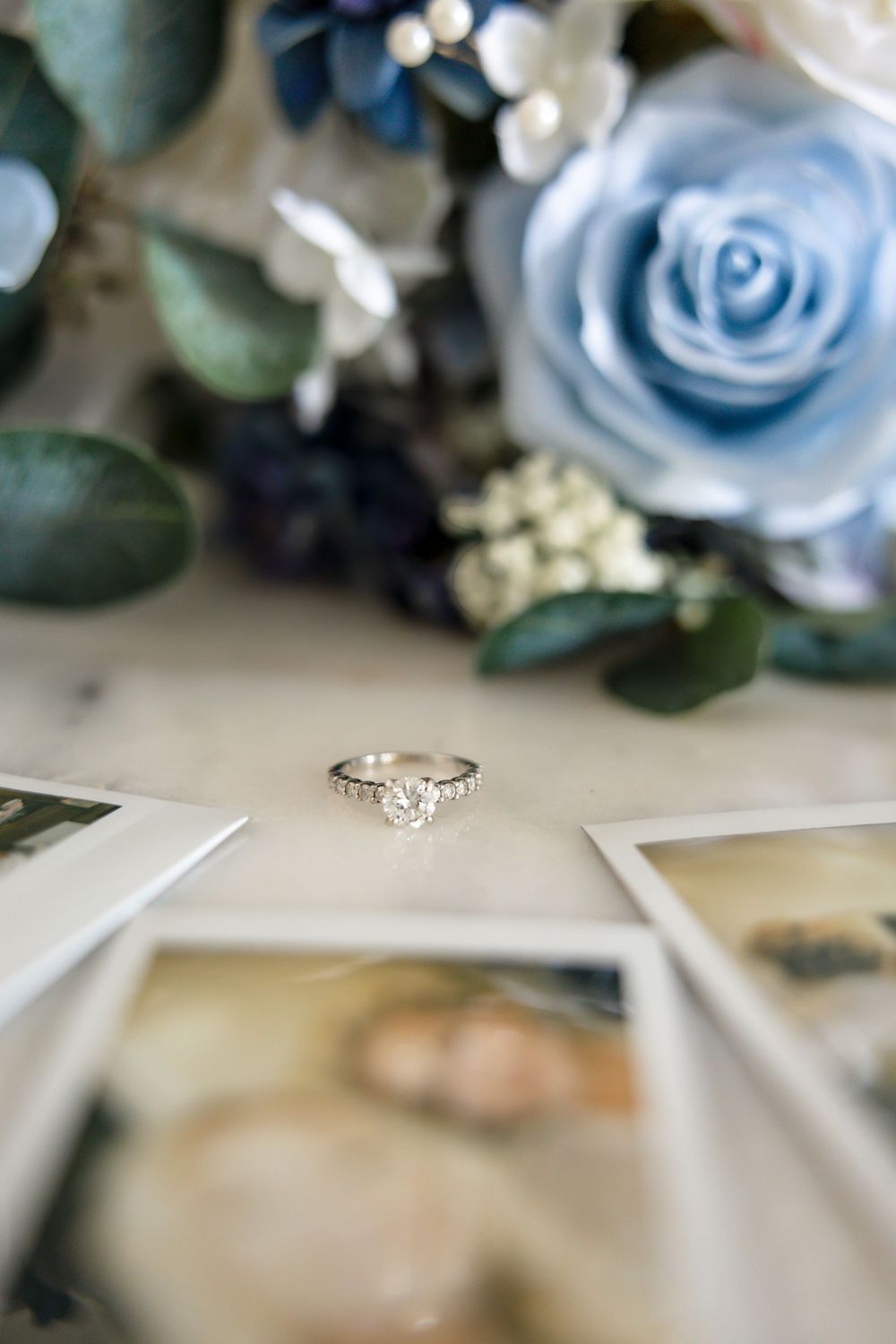 Diamond engagement ring displayed on photos beside blue roses and greenery bouquet.