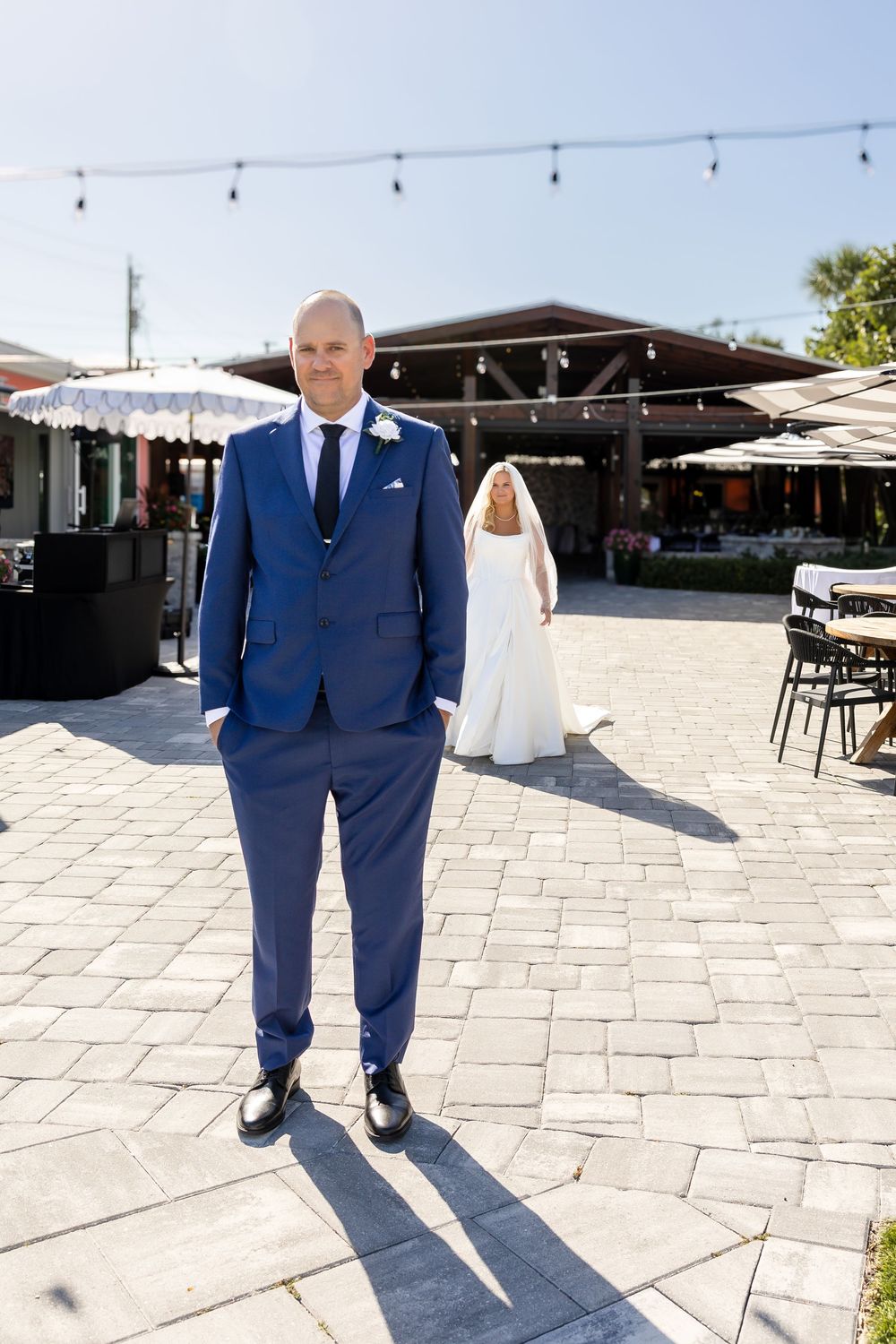 Groom in blue suit stands outdoors as bride in white dress approaches from behind.