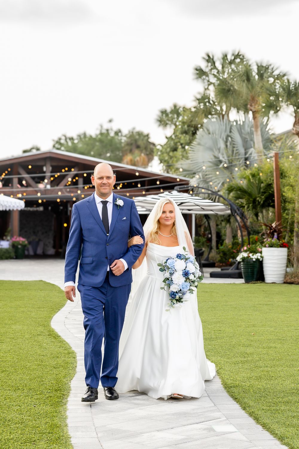 Bride and groom walking together on garden path with tropical venue in background.