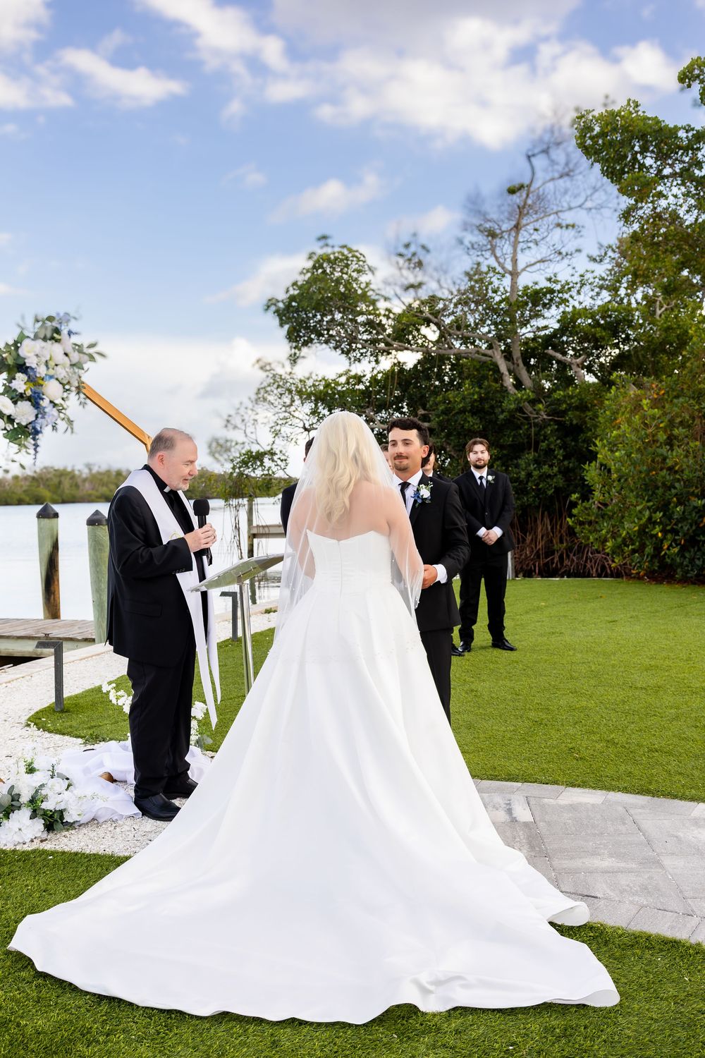 Bride in white gown with long train faces groom during outdoor waterfront ceremony.