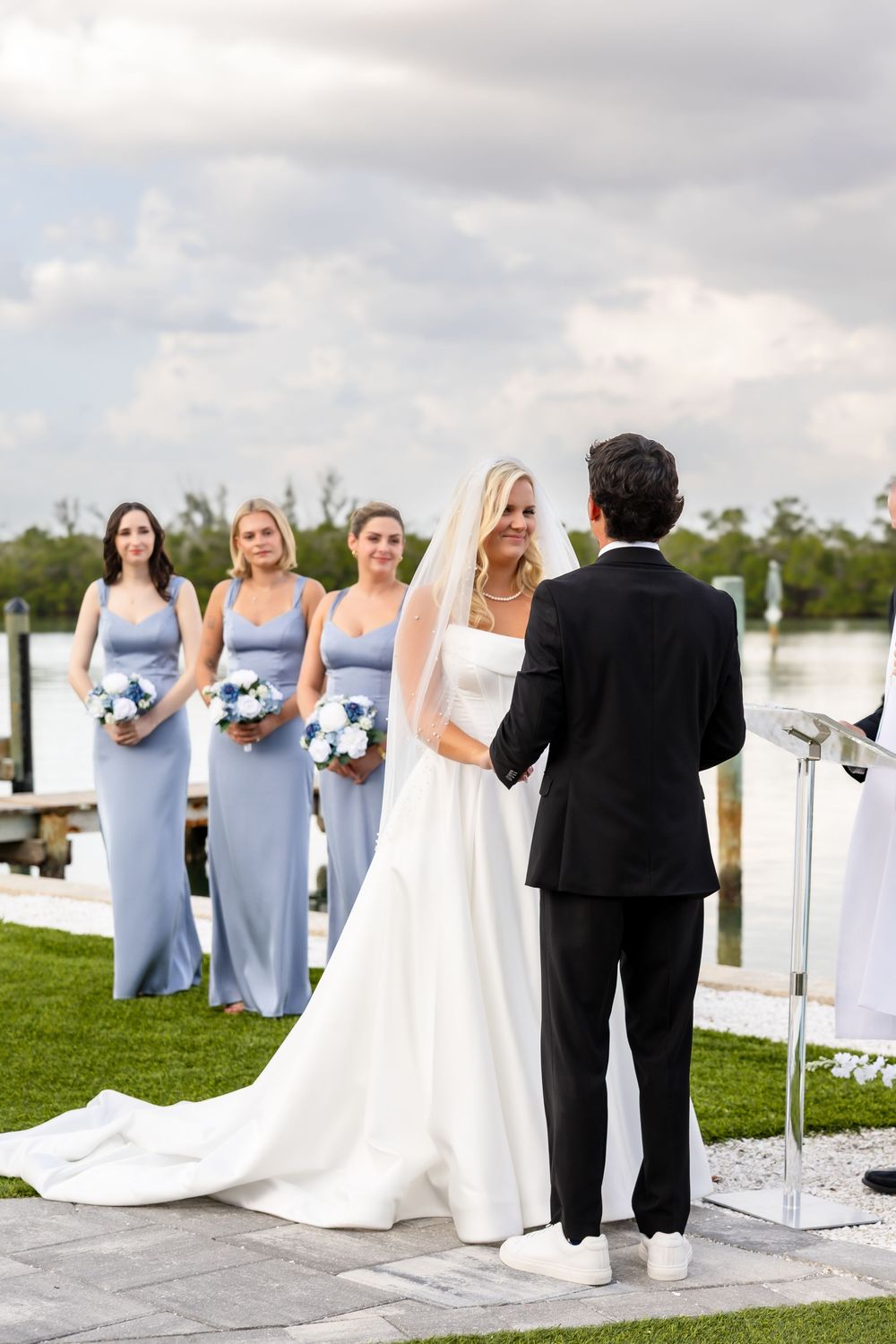 Bride and groom hold hands at altar with bridesmaids in blue dresses by waterfront.