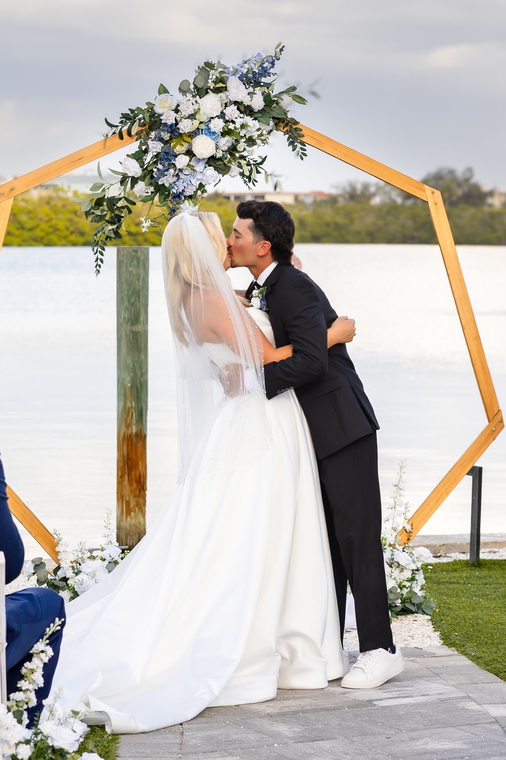 Bride and groom share first kiss under hexagonal floral arch at waterfront ceremony.
