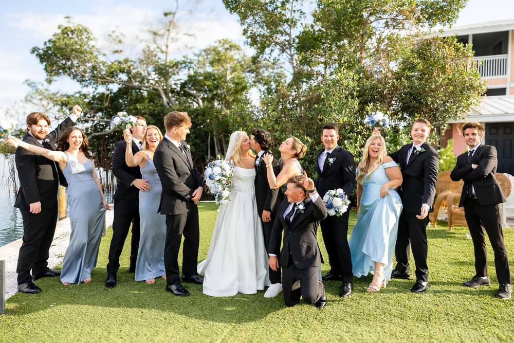 Wedding party celebrates with bride and groom kissing on lawn at outdoor venue.