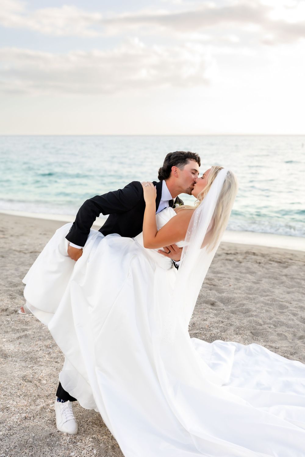 Groom dips bride for romantic kiss on sandy beach at sunset after wedding.