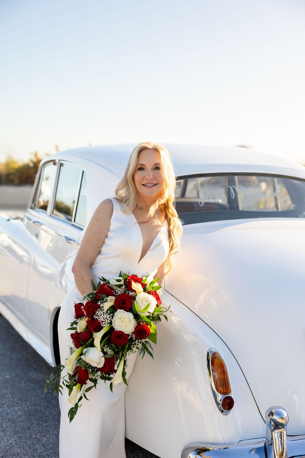 Elegant bride in white gown holding red and white bouquet leaning against vintage white car at sunset.