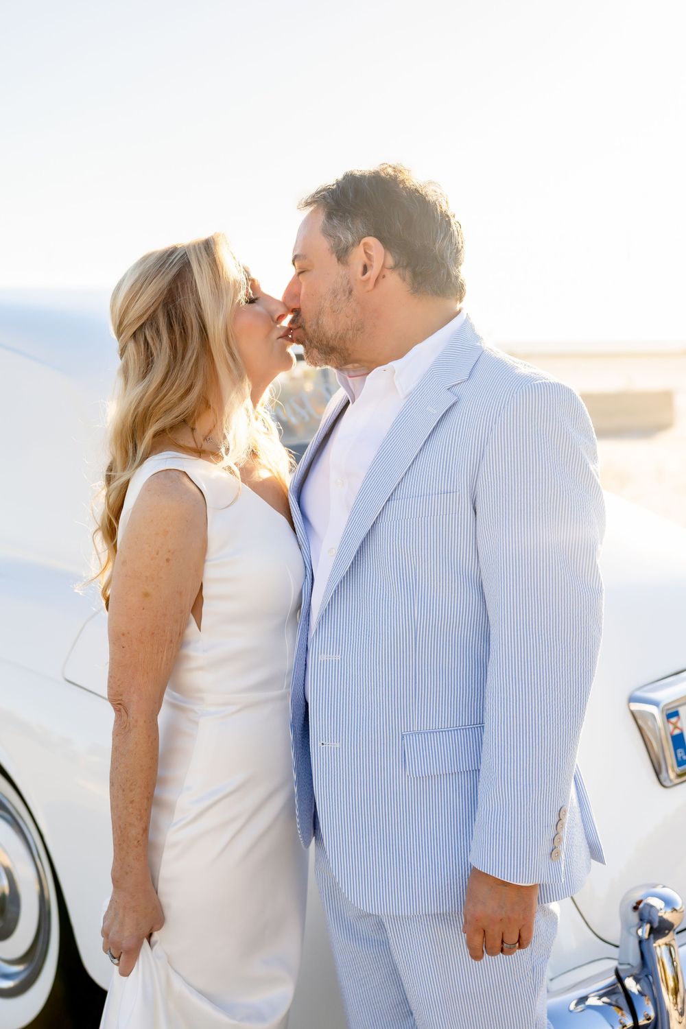 Bride and groom sharing a kiss in front of a classic white vintage car on their wedding day.