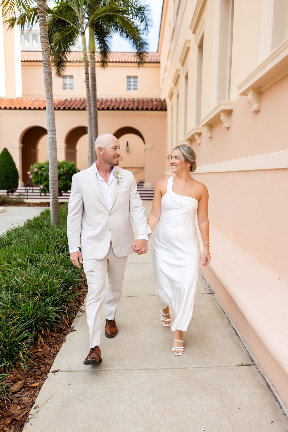 Bride and groom in white attire walk hand-in-hand along a sunlit path beside a peach Mediterranean-style building.