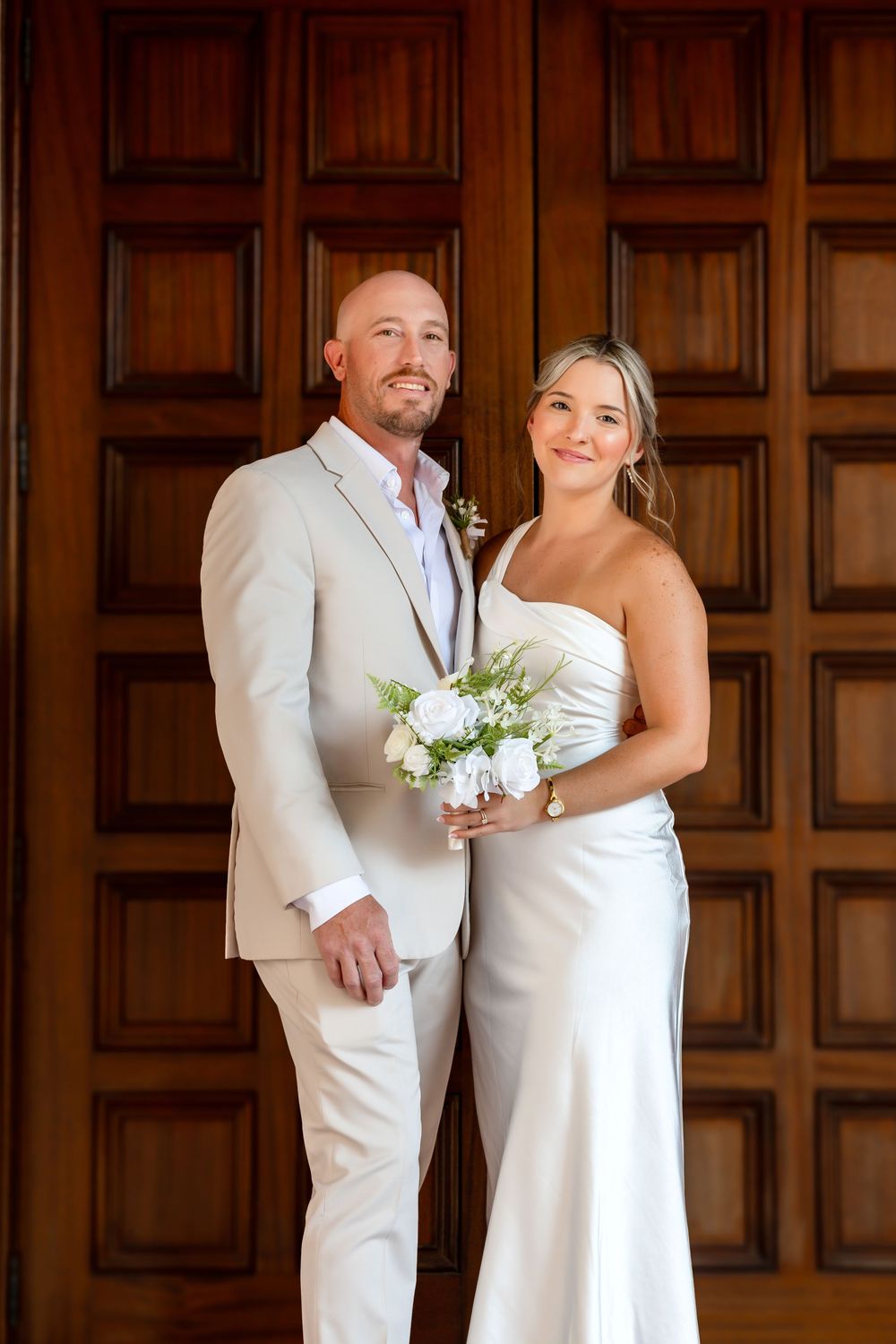 Smiling bride and groom pose before ornate wooden doors, she holds white bouquet in elegant gown, he wears beige suit.