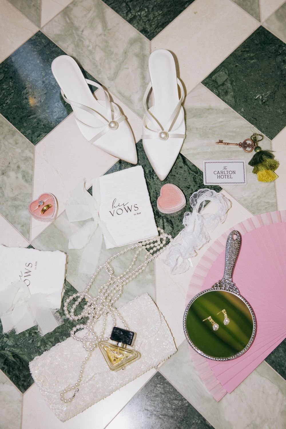 Bridal flat lay with white heels, pearl necklace, pink heart boxes, hand mirror, vows book, and Carlton Hotel key card.