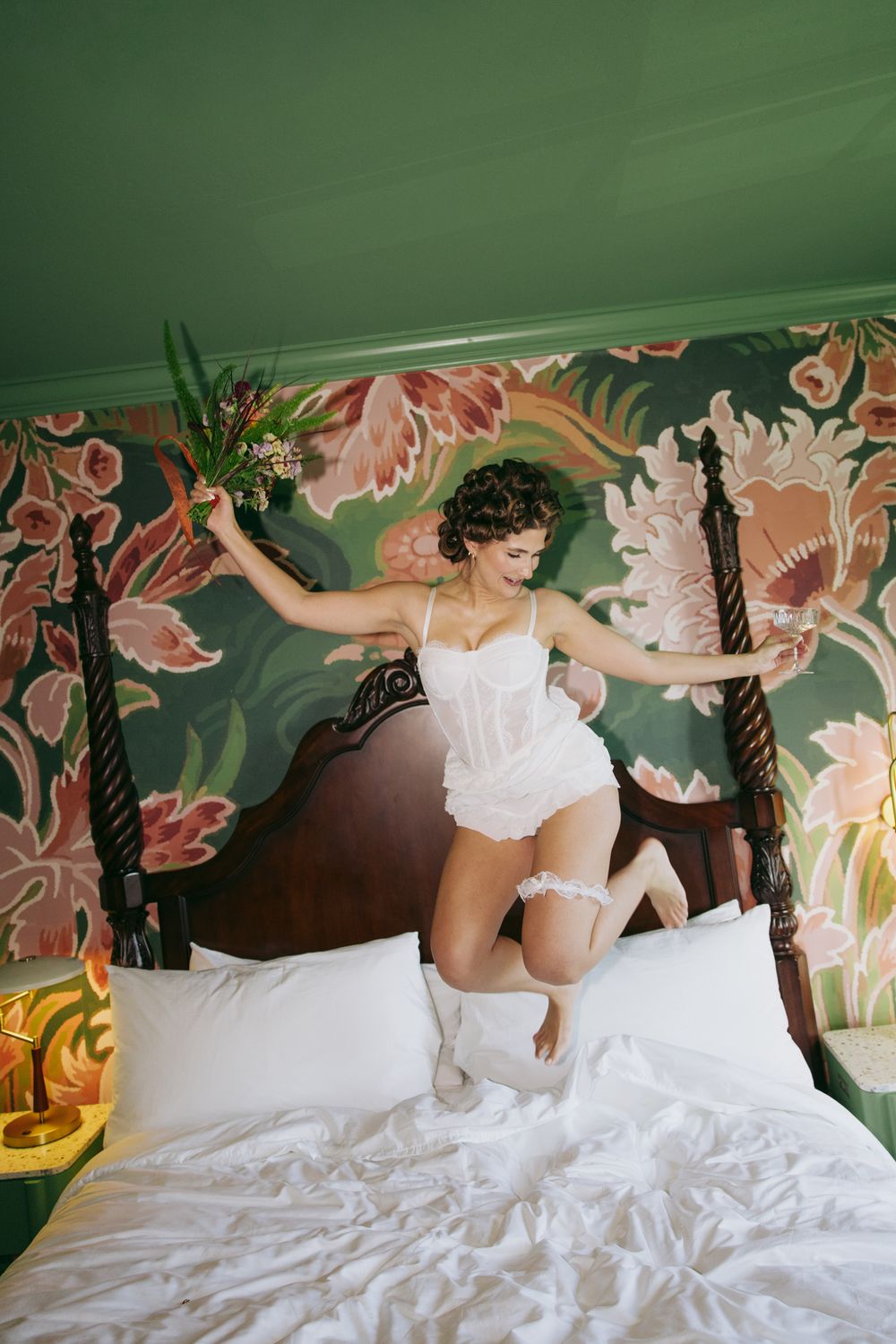 Joyful bride in white corset lingerie jumping on bed in floral wallpaper hotel room holding flowers.