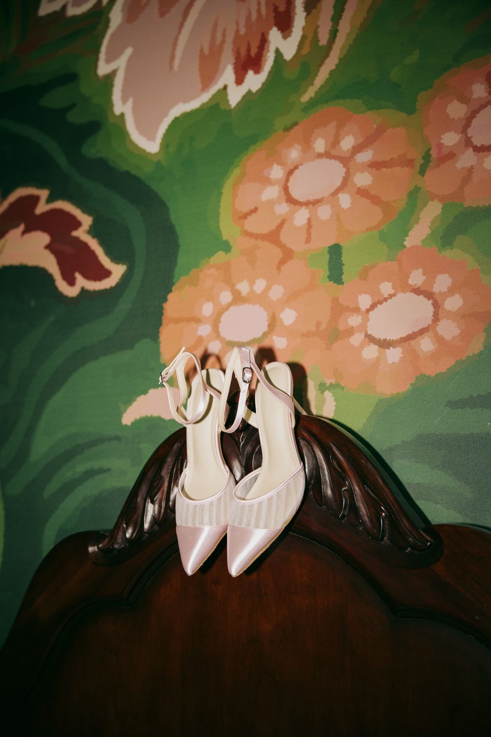 White bridal heels with ankle straps displayed on dark wood chair against vibrant floral green wallpaper.
