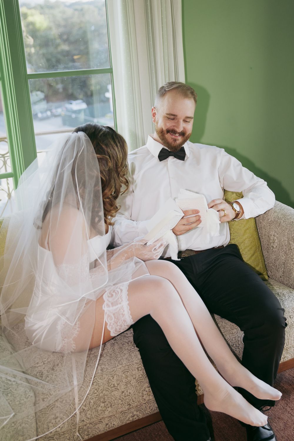 Bride and groom sitting together on a couch, smiling and reading notes in a sunlit room on their wedding day.