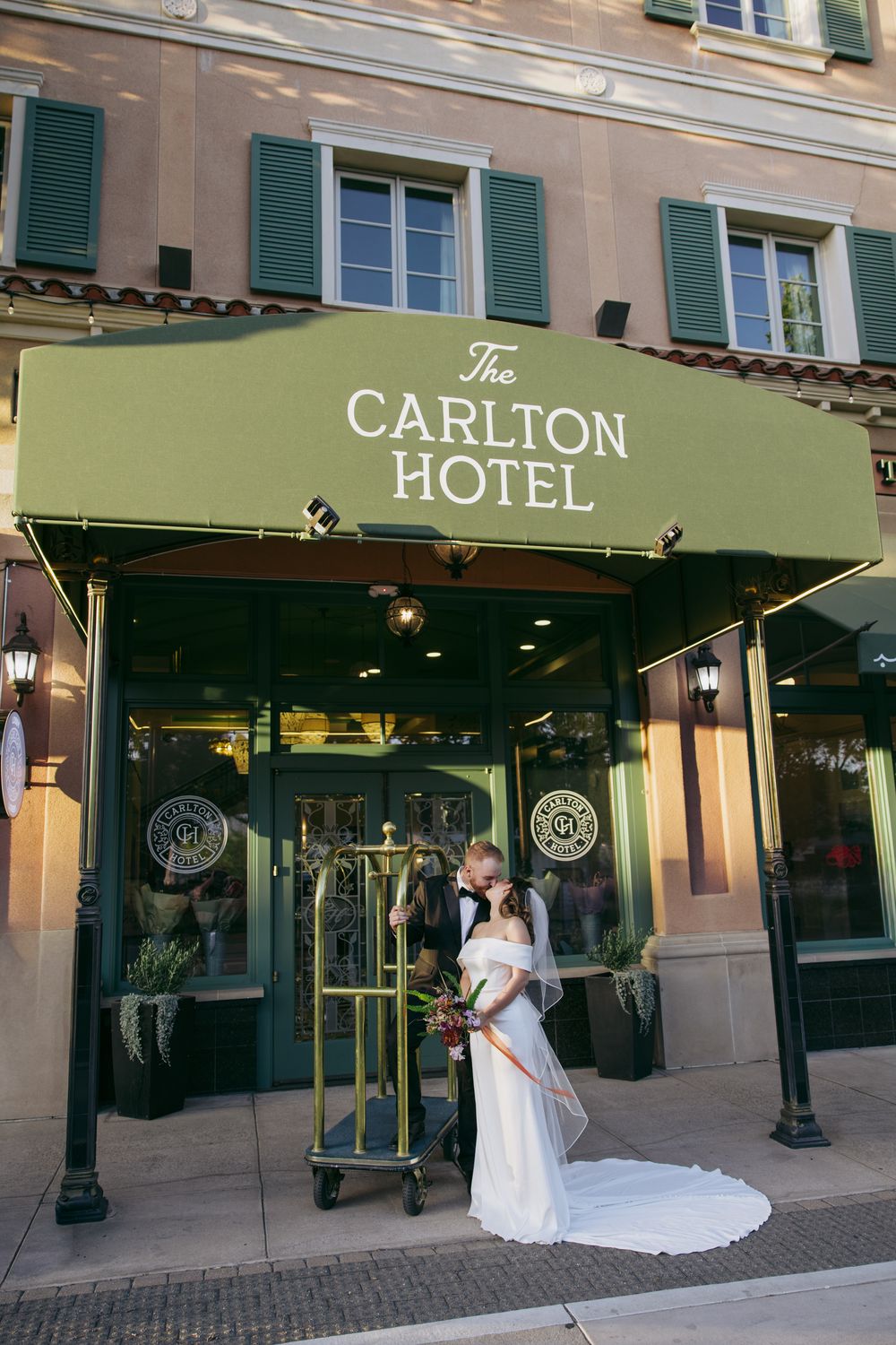 Bride and groom kissing under green Carlton Hotel awning entrance on their wedding day.