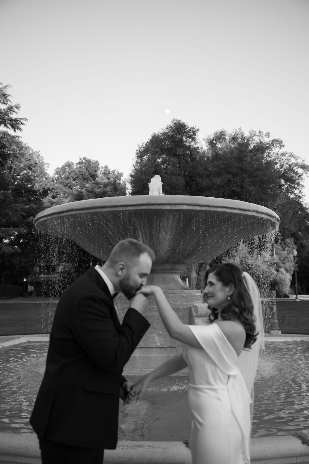 Black and white photo of a groom kissing his bride's hand near a fountain outdoors.