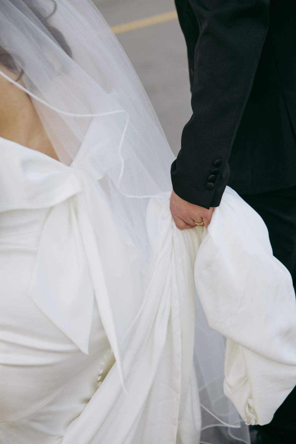 Bride in white gown with bow detail holds groom's hand in black suit, wedding dress train flowing gracefully.