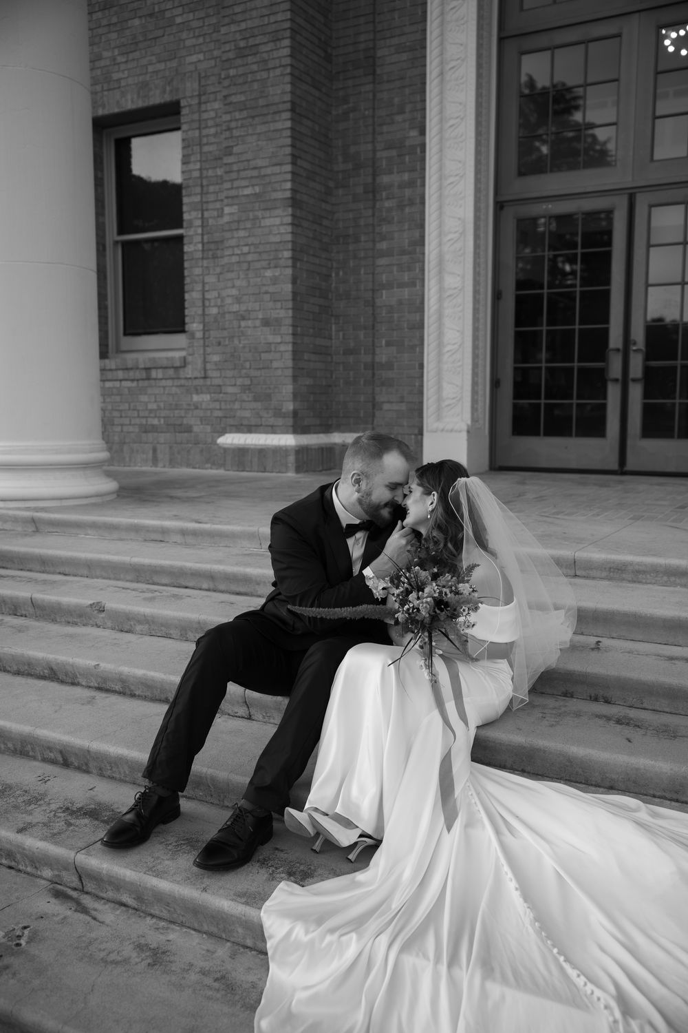 Black and white photo of bride and groom kissing on steps outside a brick building on their wedding day.