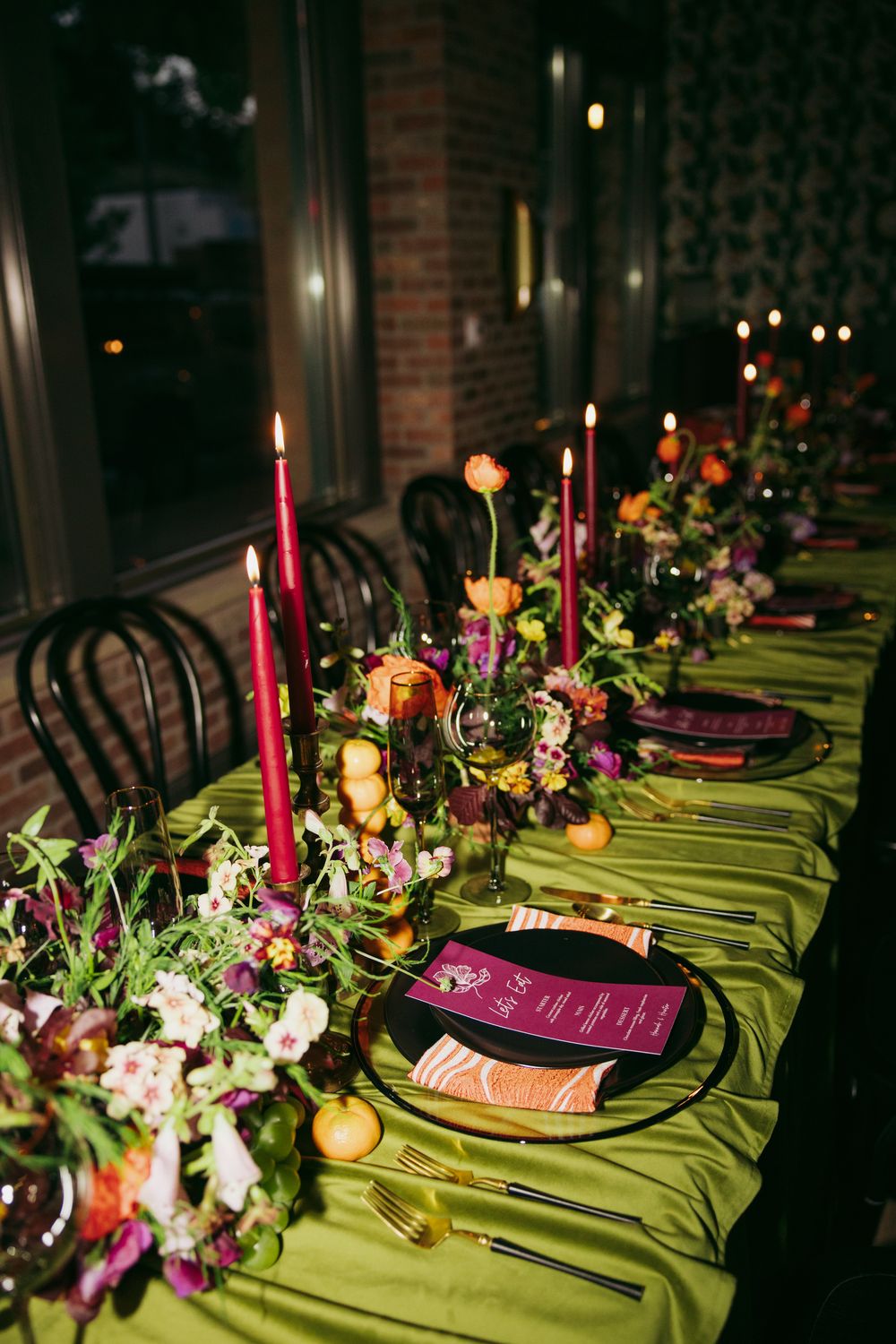 Elegant wedding reception table with green satin cloth, magenta candles, gold cutlery, black plates, and colorful floral centerpieces.