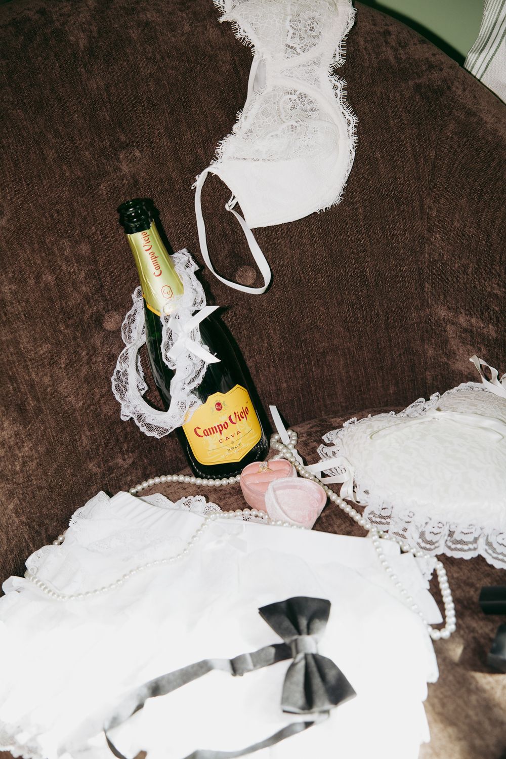 Champagne bottle with Campo Viejo label surrounded by white lace lingerie and pearl necklace on brown velvet surface.
