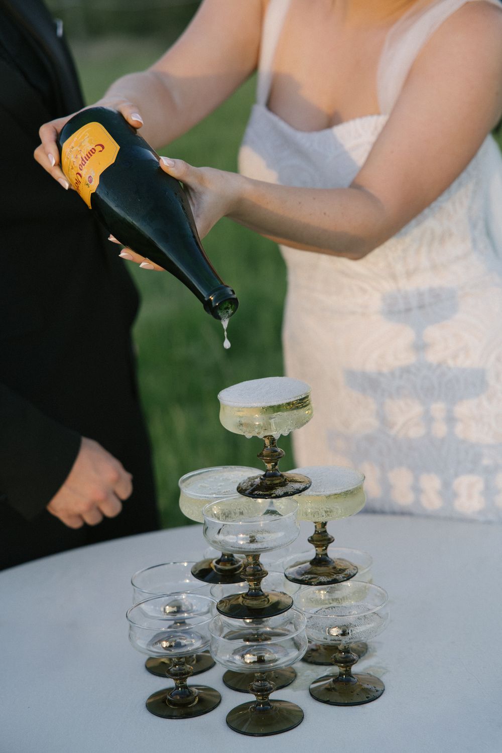 Bride and groom pouring champagne tower with dark coupe glasses at outdoor wedding reception.