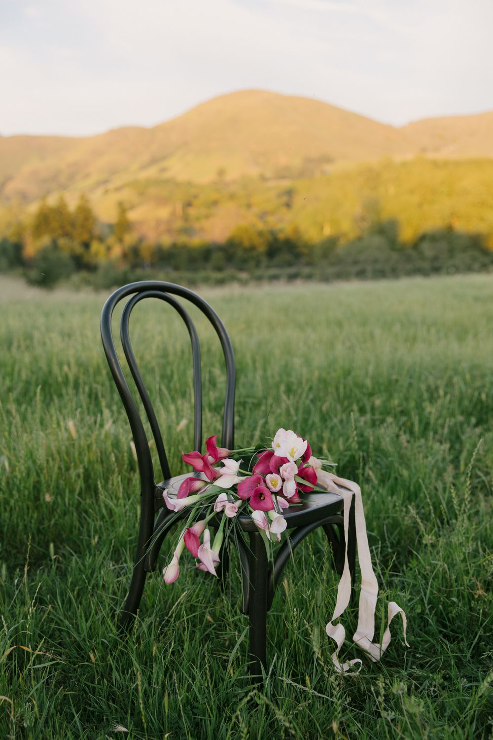 Black bistro chair holding pink and white floral bouquet with ribbons in green meadow with mountain backdrop at sunset.