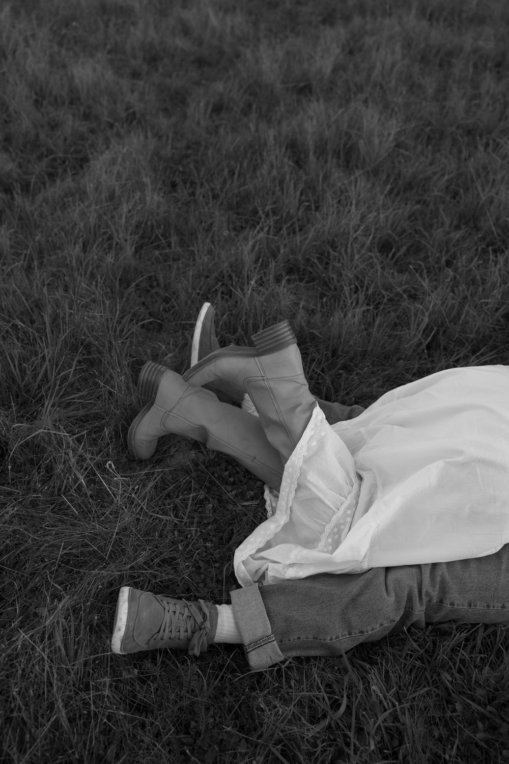 Black and white photo of two people lying on grass, wearing boots and a white skirt.