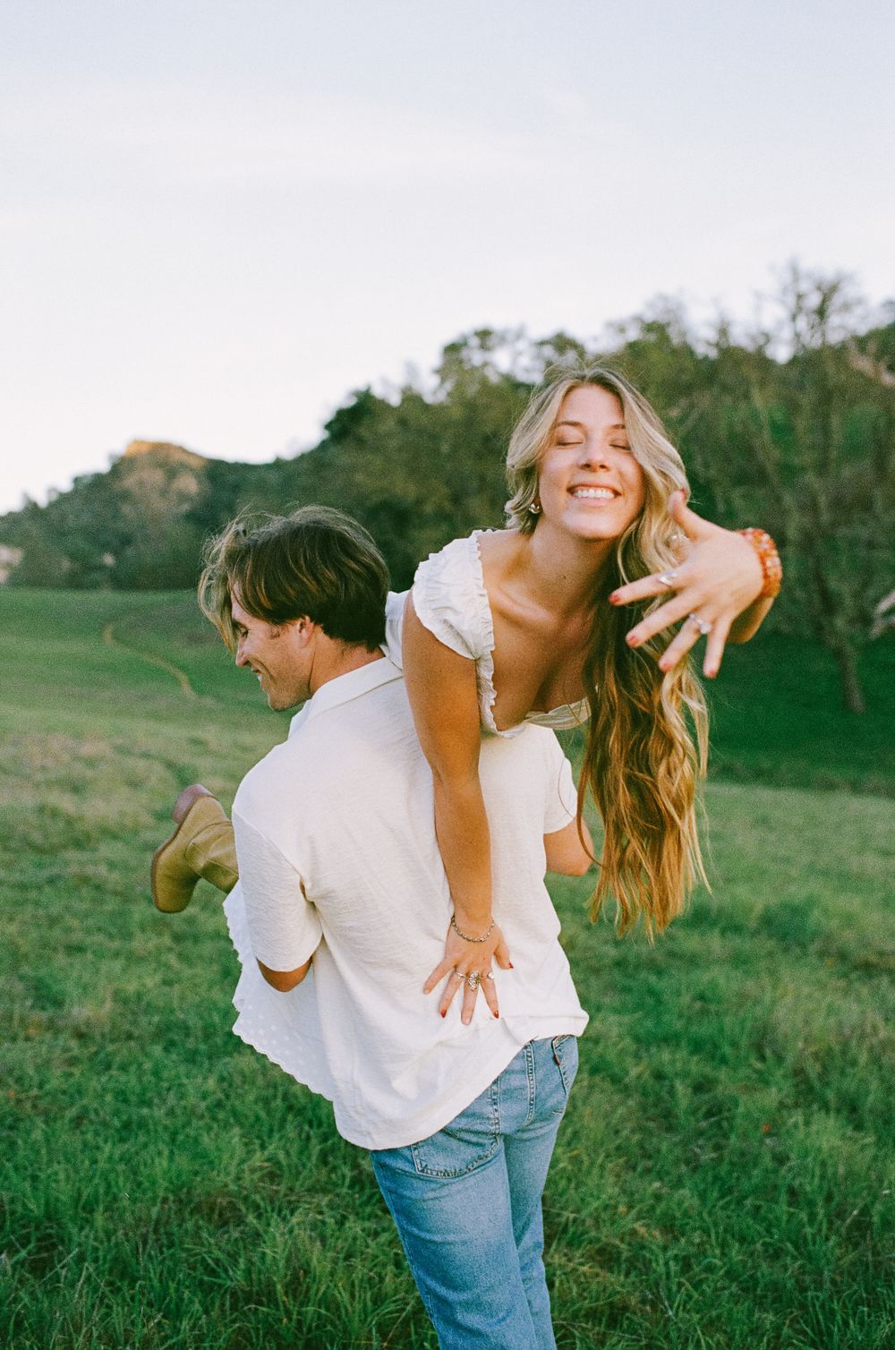 A joyful couple in a green field, man giving woman a piggyback ride, both laughing, wearing white tops and jeans.