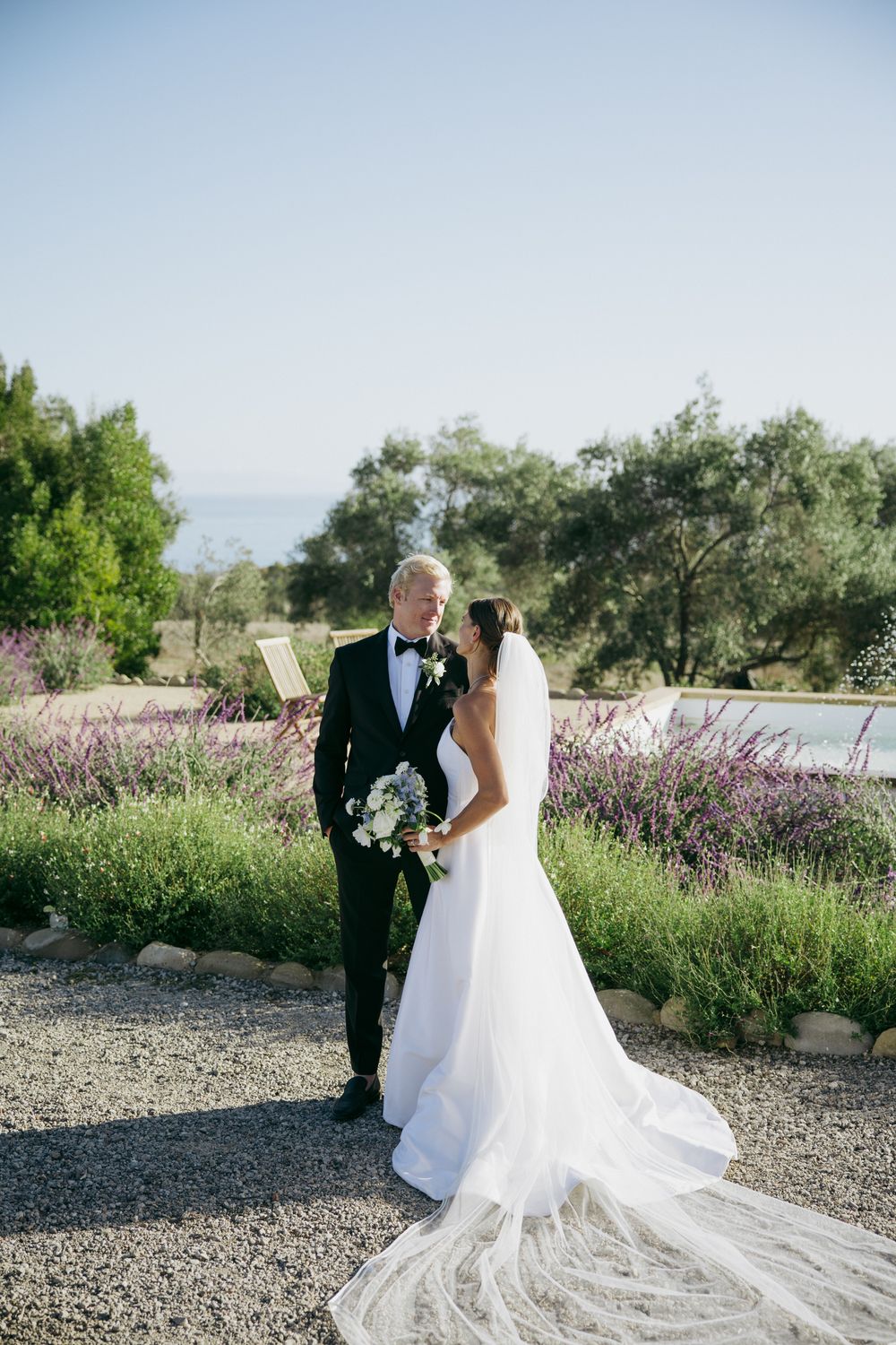 A couple in wedding attire poses in front of blooming lavender fields on a bright sunny day.