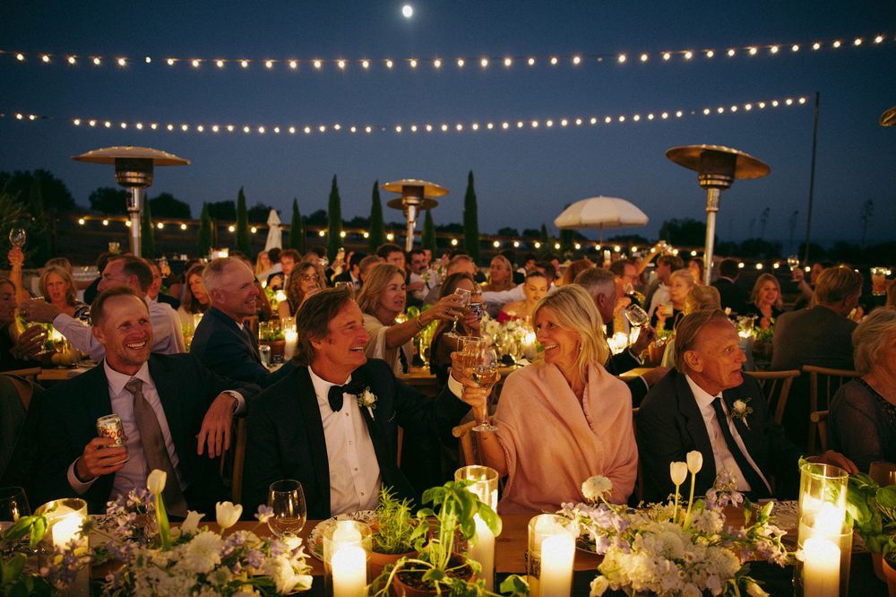 String lights illuminate an elegant outdoor evening reception with guests seated at long tables for dinner.