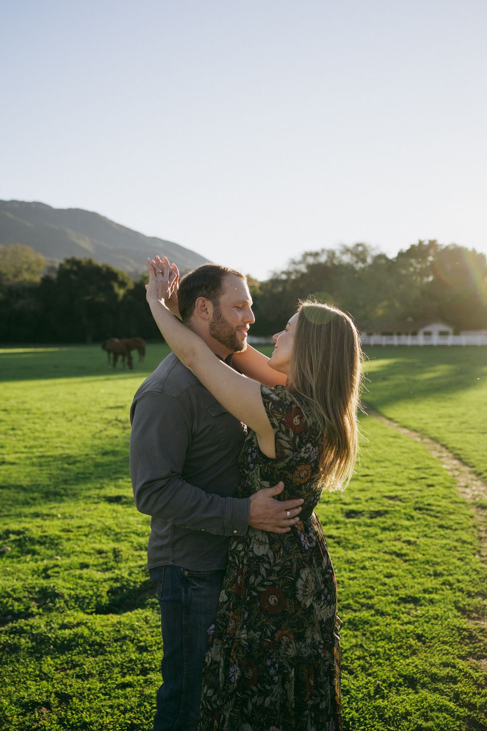 A couple embraces and kisses in a sunlit green field with mountains in the background during golden hour.