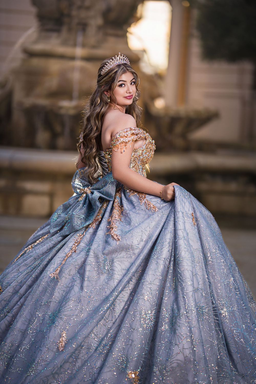Young woman in a sparkling blue ball gown with gold details and tiara poses elegantly near a fountain.