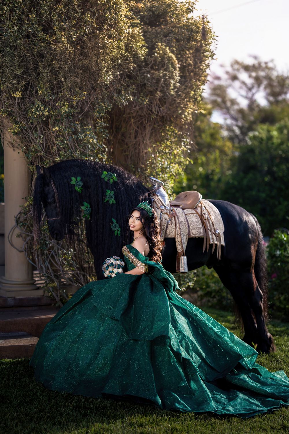 Young woman in emerald green ball gown seated beside a black horse with saddle outdoors.