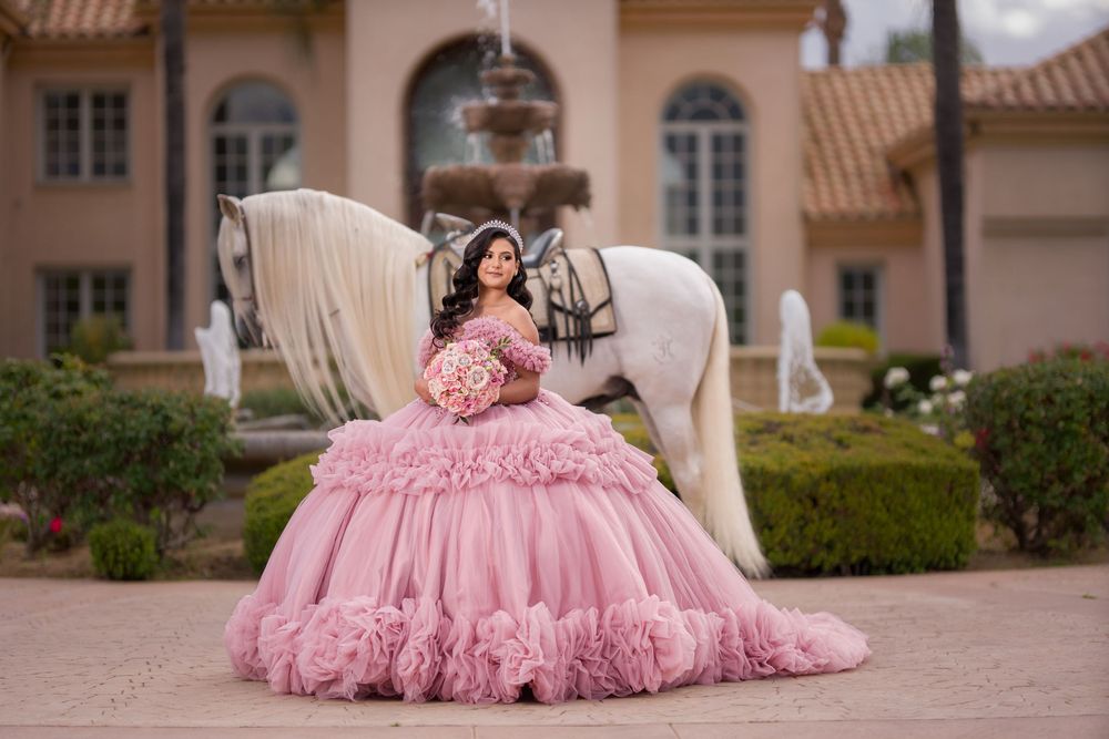 Young woman in pink ball gown poses with white horse at elegant estate fountain for quinceañera photos.