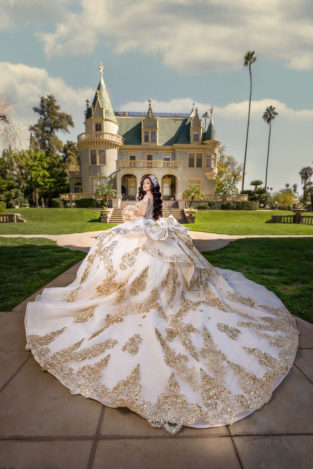 Woman in ornate white and gold ball gown poses before a grand castle-like mansion with lush green lawns.