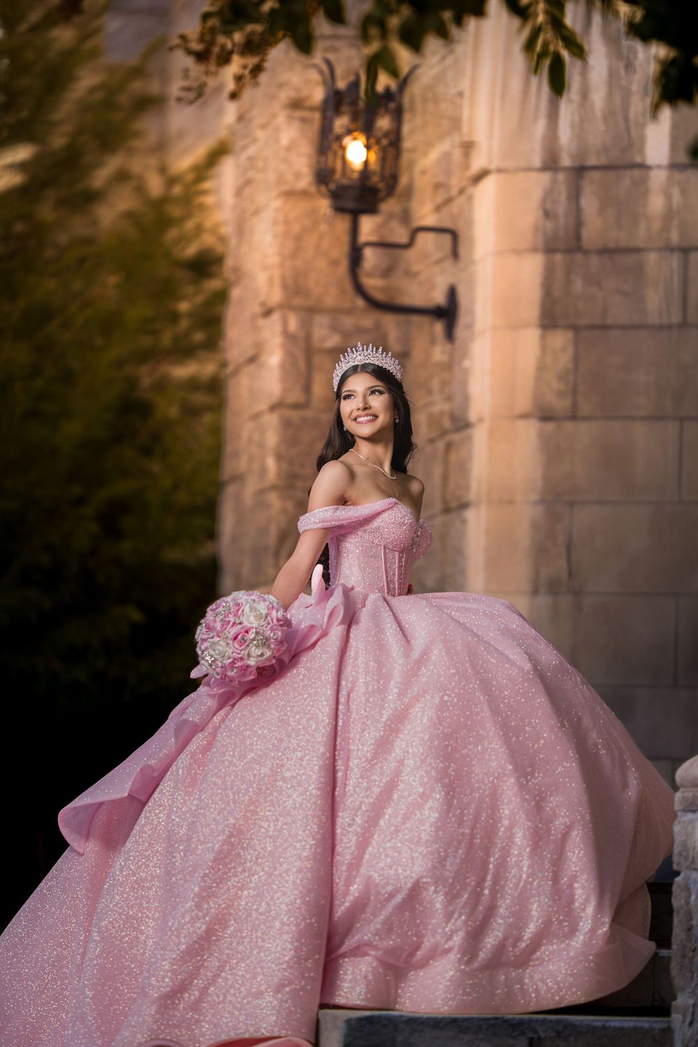 A sparkling pink quinceañera ball gown photographed in an elegant outdoor setting with stone archways and warm lighting.