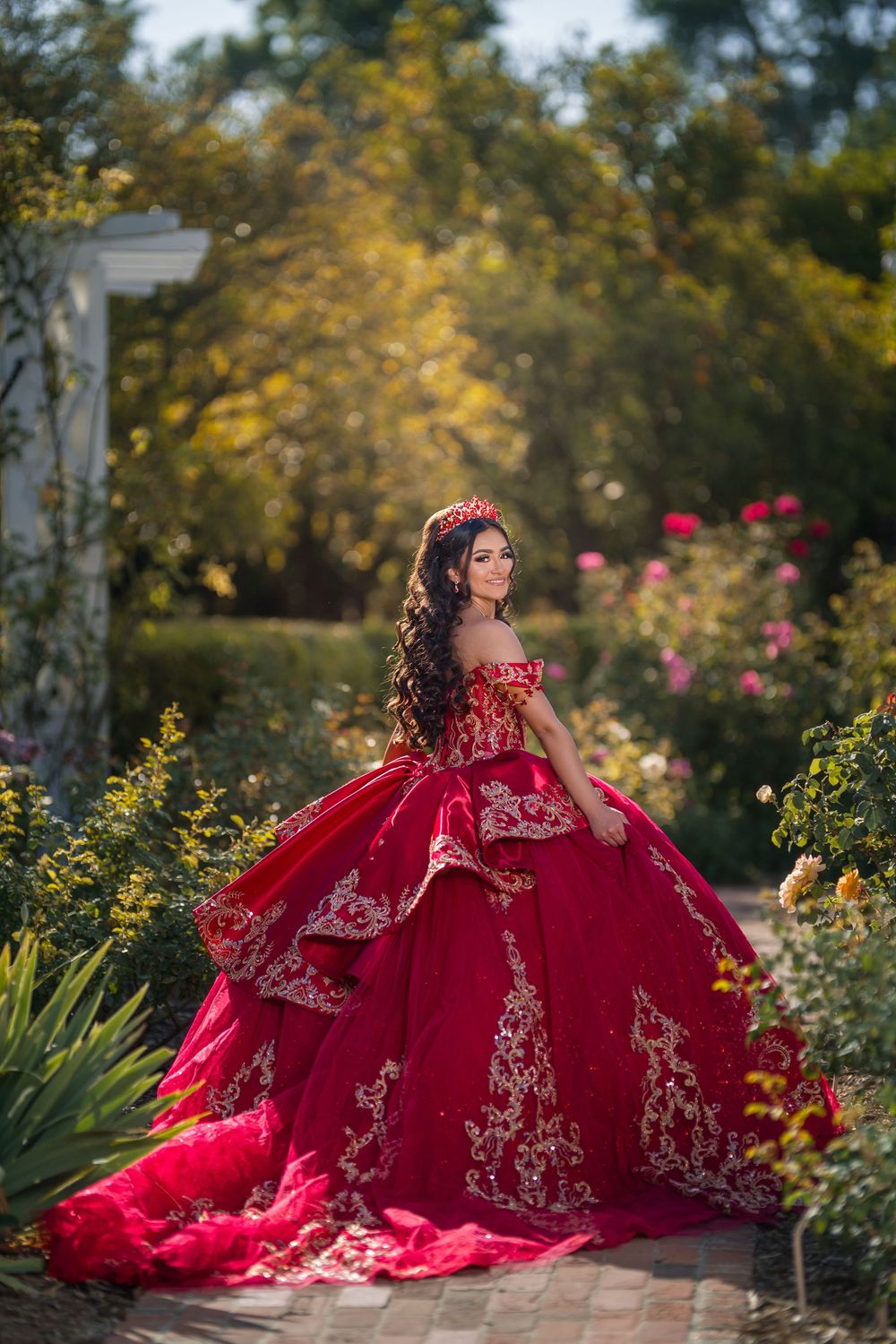 Young woman in red quinceañera ball gown with gold embroidery poses in a lush garden surrounded by colorful roses.