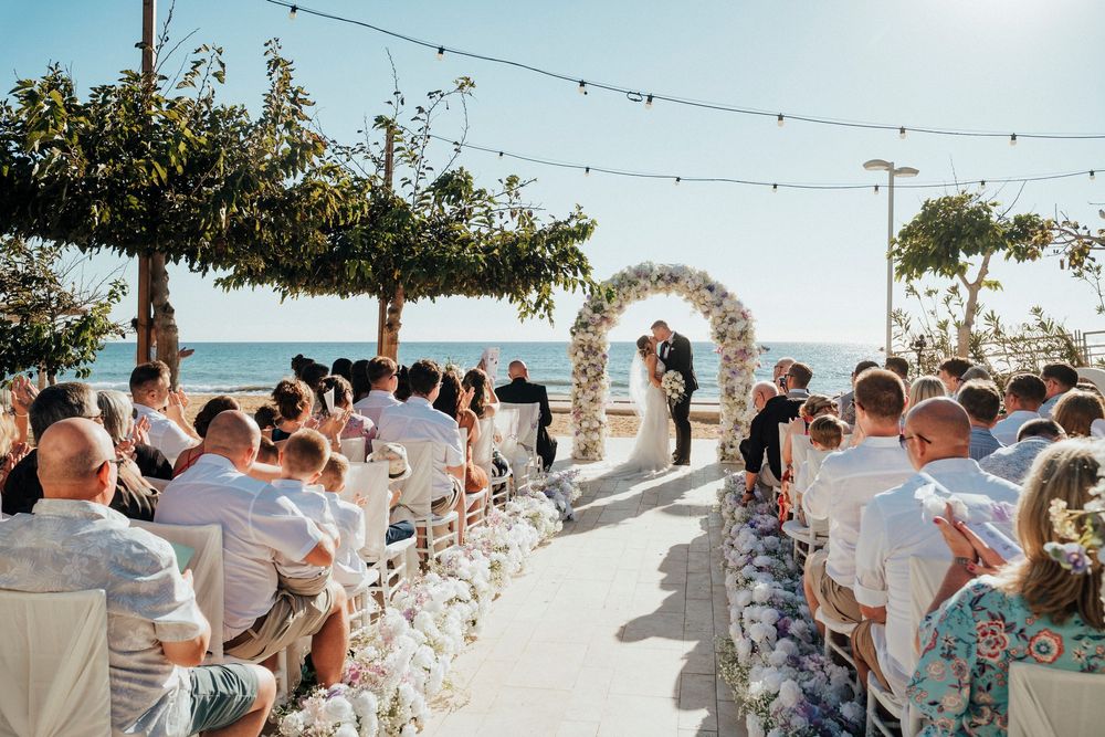 Outdoor beach wedding ceremony with string lights and white decor along a sandy path leading to the ocean.