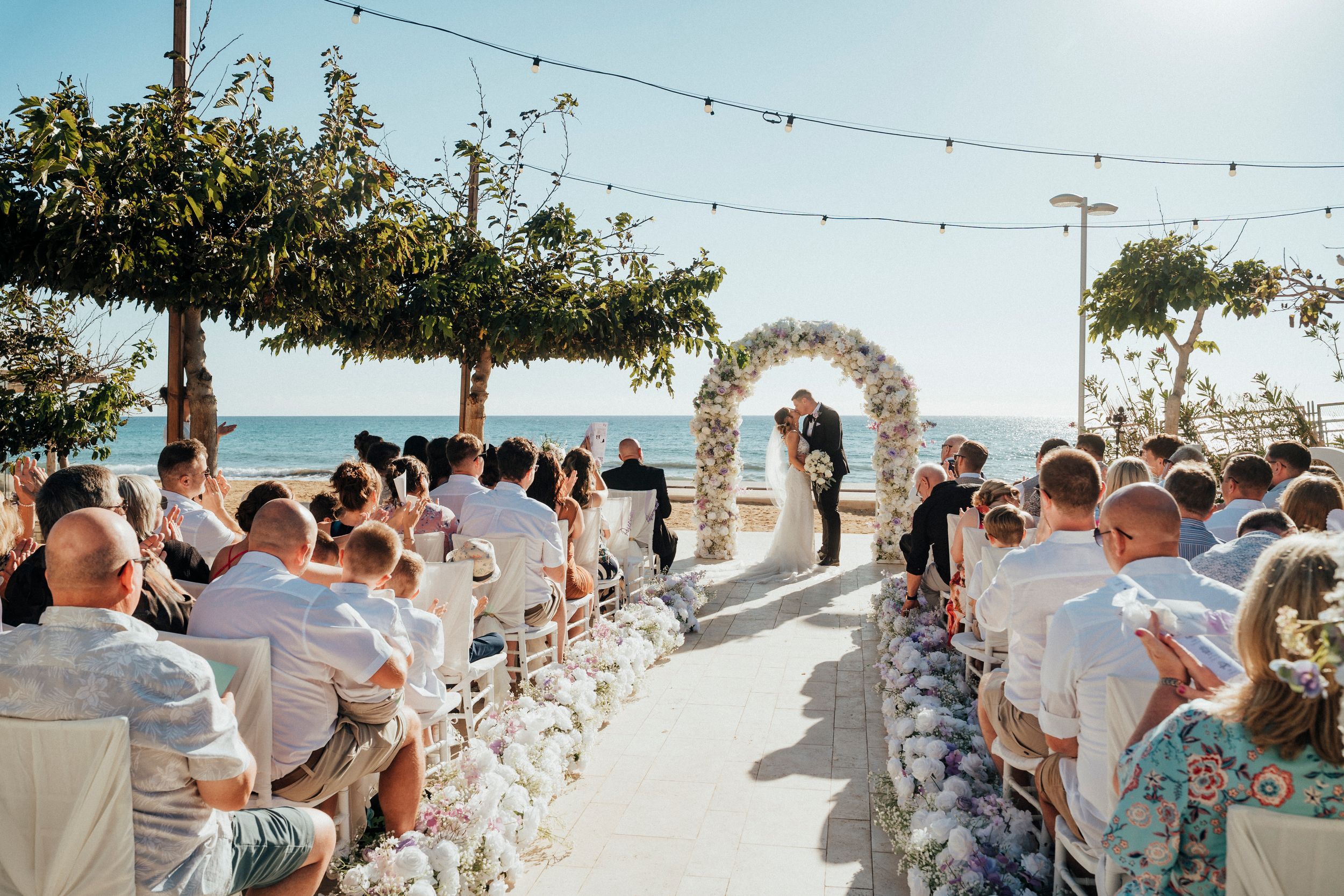 Outdoor beach wedding ceremony with string lights and white decor along a sandy path leading to the ocean.