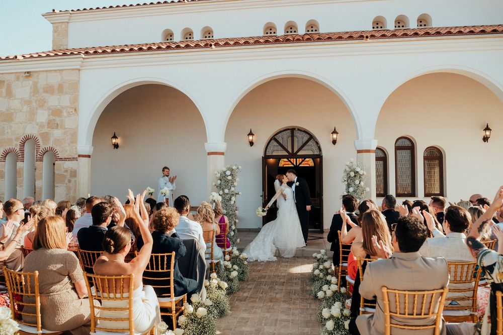 Wedding guests raise their arms in celebration during an outdoor reception under white arched columns at sunset.