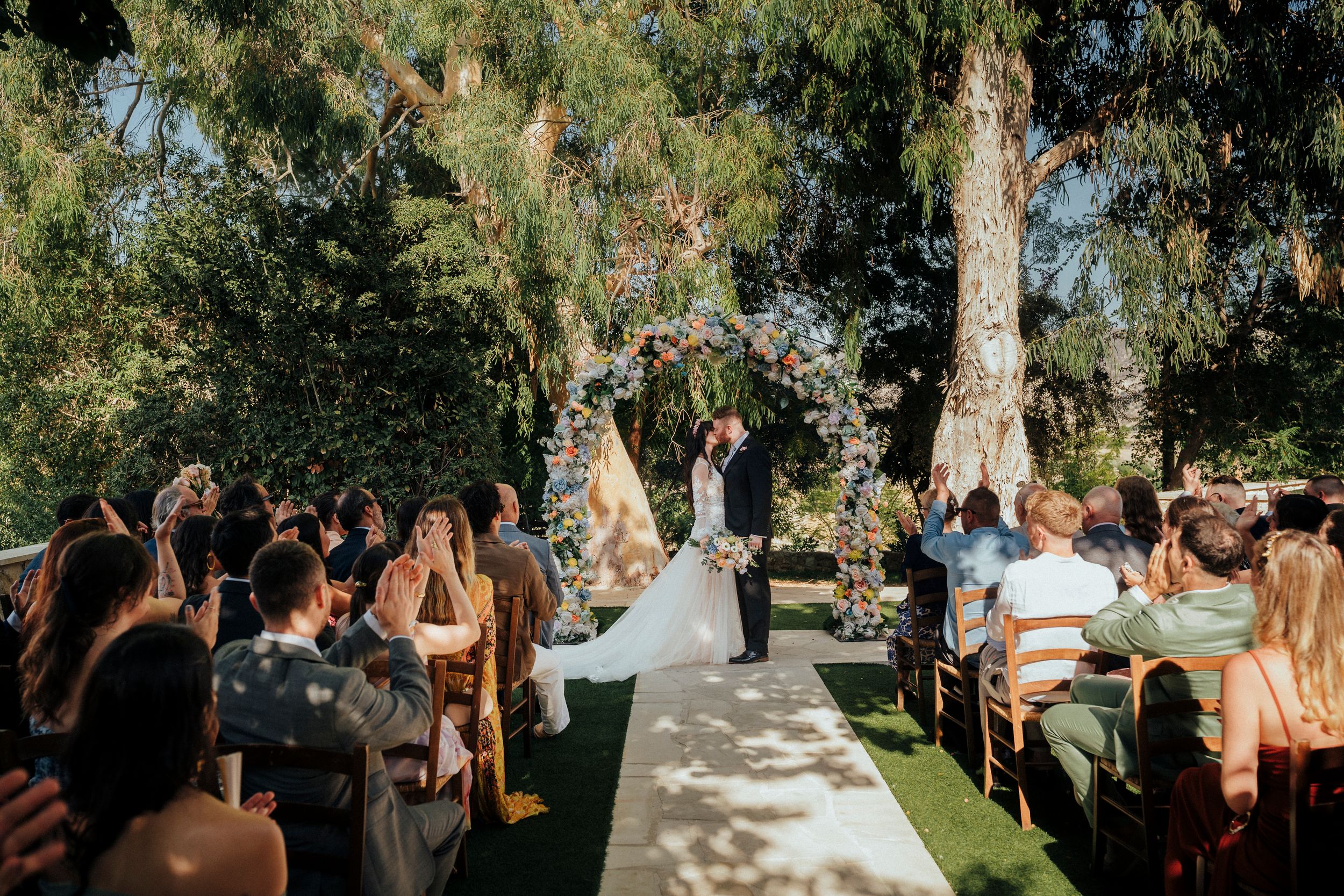 Wedding guests gather outdoors under tall trees during a sunny garden ceremony celebration.