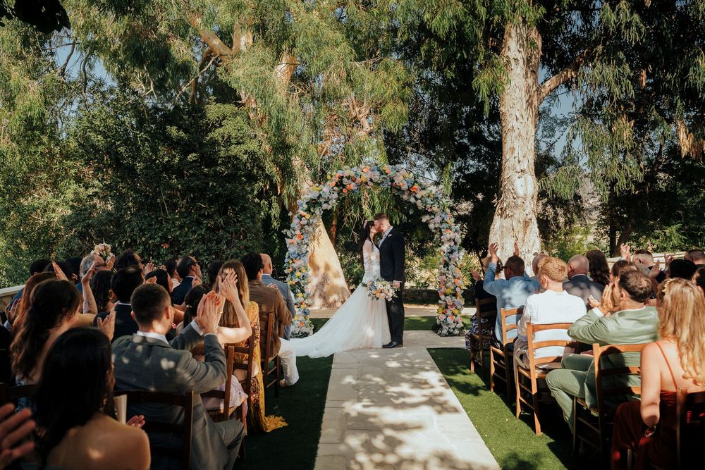 Wedding guests gather outdoors under tall trees during a sunny garden ceremony celebration.