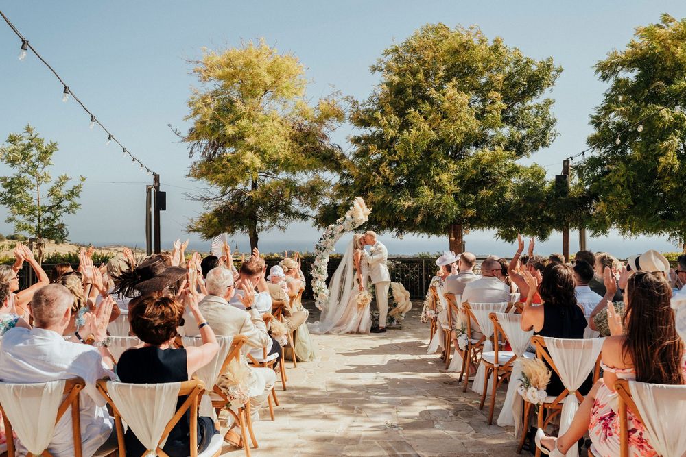 Wedding guests dance and celebrate on an outdoor patio surrounded by trees during a sunny afternoon reception.