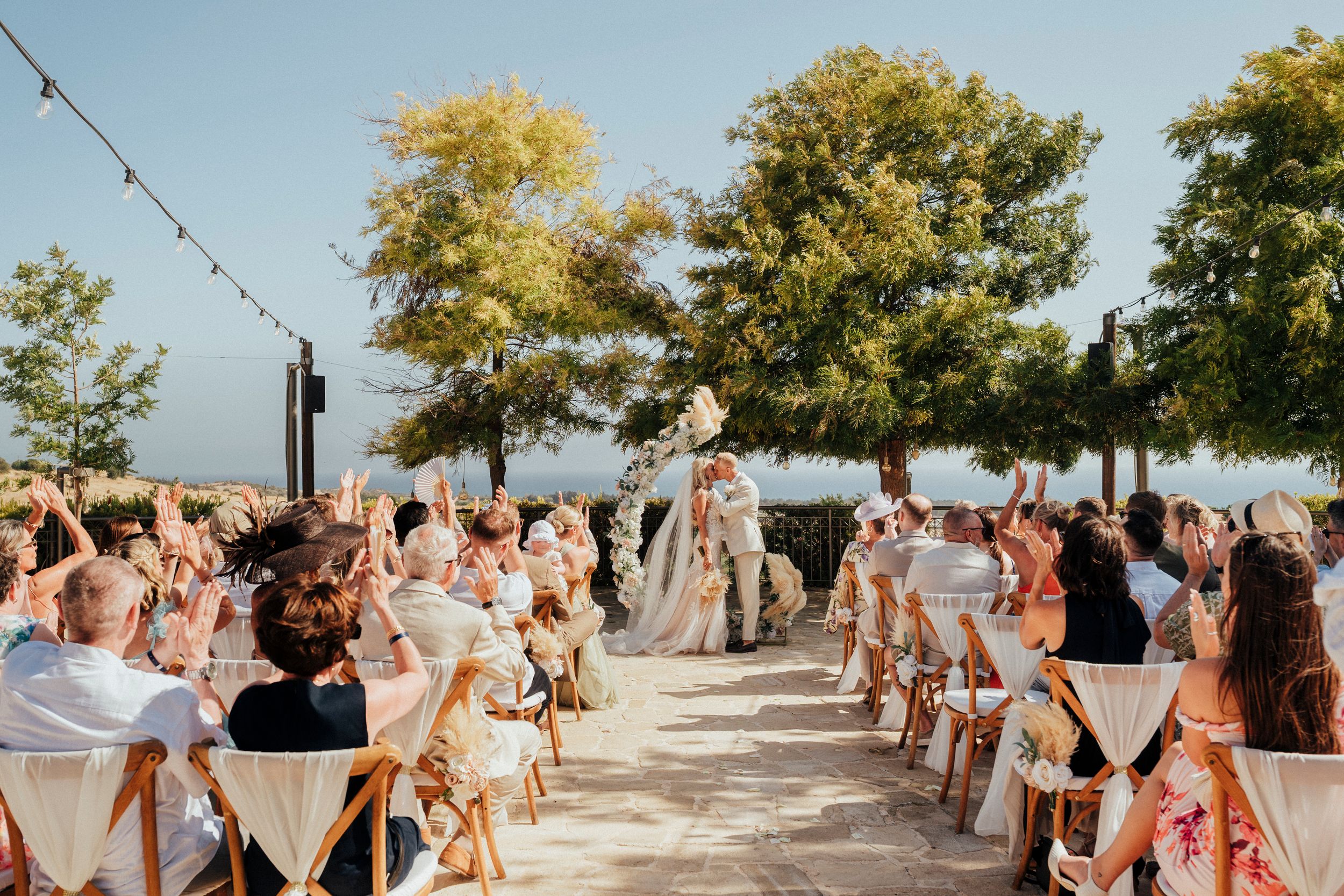 Wedding guests dance and celebrate on an outdoor patio surrounded by trees during a sunny afternoon reception.