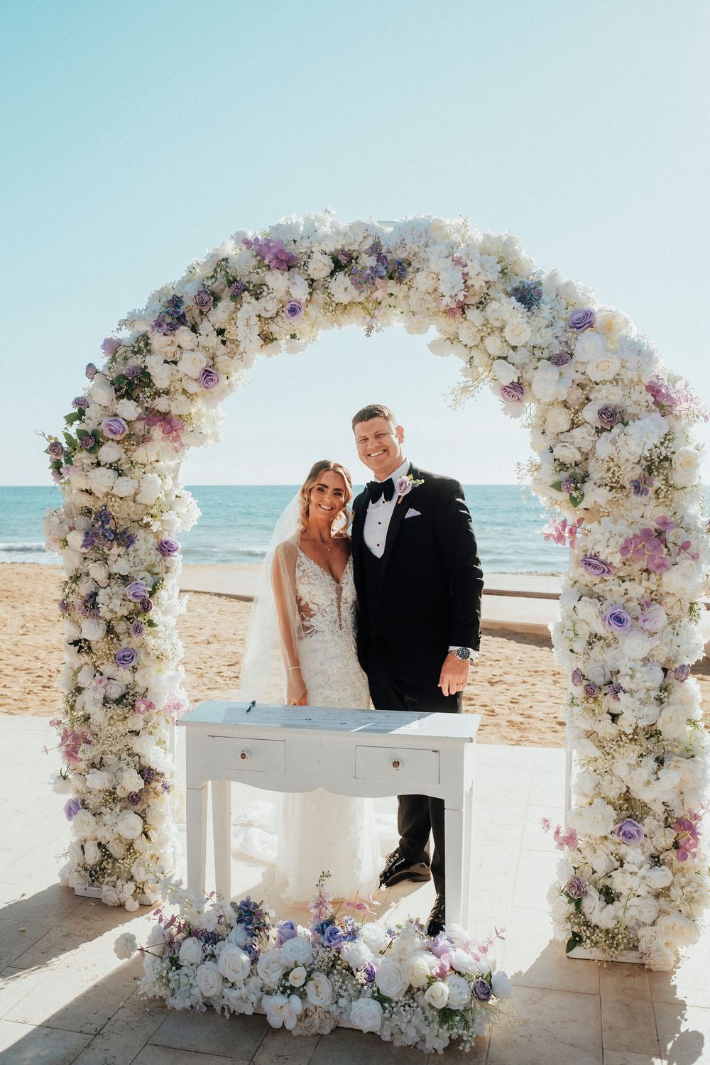 A romantic beach wedding ceremony setup with a white floral arch and white piano against the ocean backdrop.
